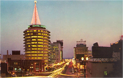 A busy city street at night (just as the sun is setting) with a tall cylinder-shaped building. At the top of the building, there is a red-lighted christmas tree, topped with a golden star. Along the rim of the upper-portion of the building are the words "CAPITOL RECORDS" written several times.