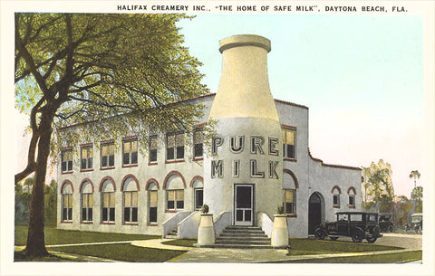 Vintage image of a two-story building, shot in daylight,  with a gigantic over two-story high entrance in the shape of a milk bottle which has the words at top:  "Pure Milk" written on two lines in all cap neon letters and a black antique car parked off to the rjght side. On the left side there is a large tree.