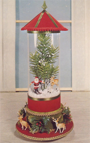 "Santa Clause is skiing on a pile of rounded snow alongside a reindeer and in front of a pine tree that is covered in snow.  At the base of the Christmas decoration, there are several reindeer and holly plants along the circumference of the circular  base that is trimmed in a gold pattern. "